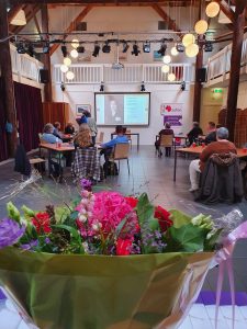 Zaal met mensen die luisteren naar een spreker met op de voorgrond bloemen
