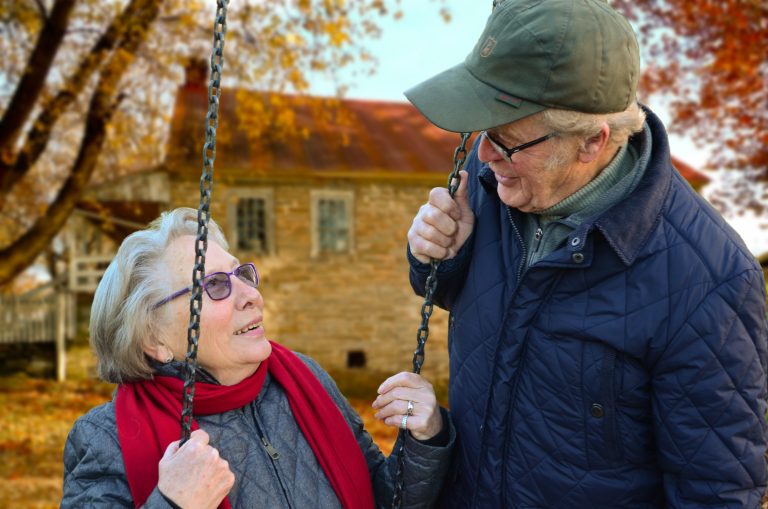 Ouder echtpaar kijkt naar elkaar, vrouw zit op schommel.