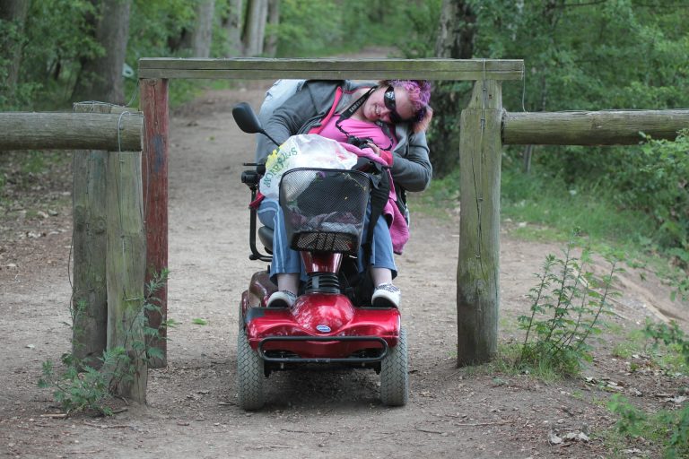 vrouw in een scootmobiel die moet bukken om door een hekje te komen